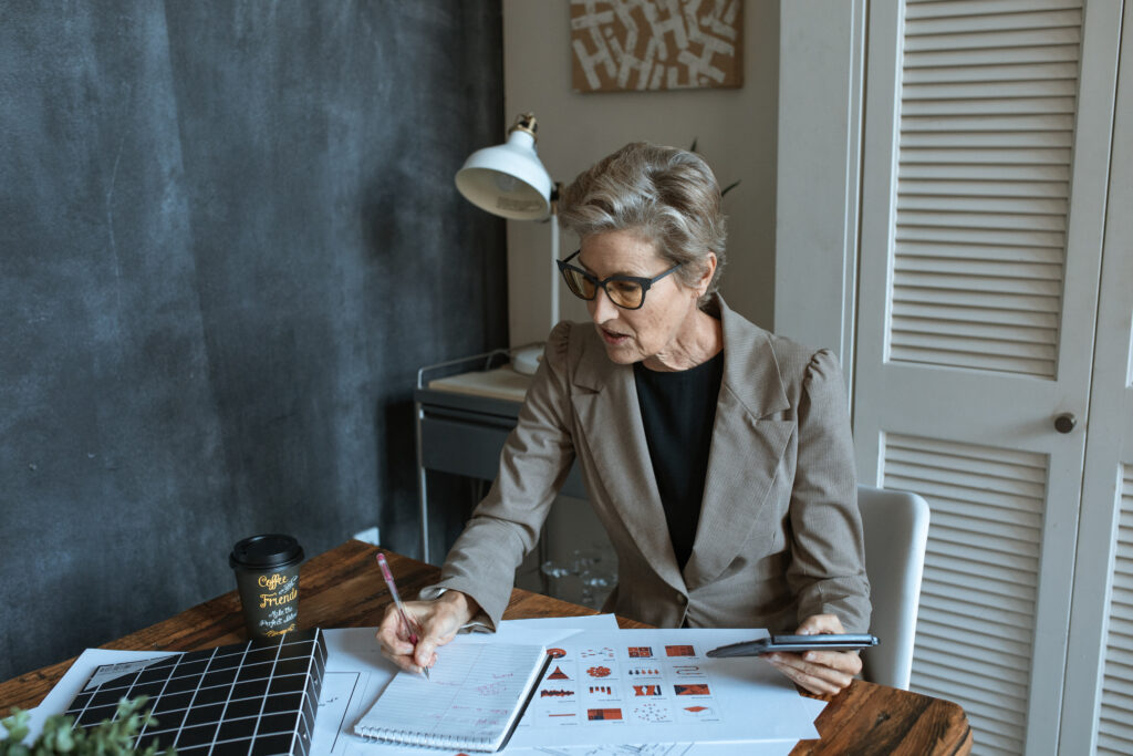 From above of concentrated senior female manager in formal outfit and eyeglasses taking notes in planner while working at table with various documents and takeaway coffee in cozy office