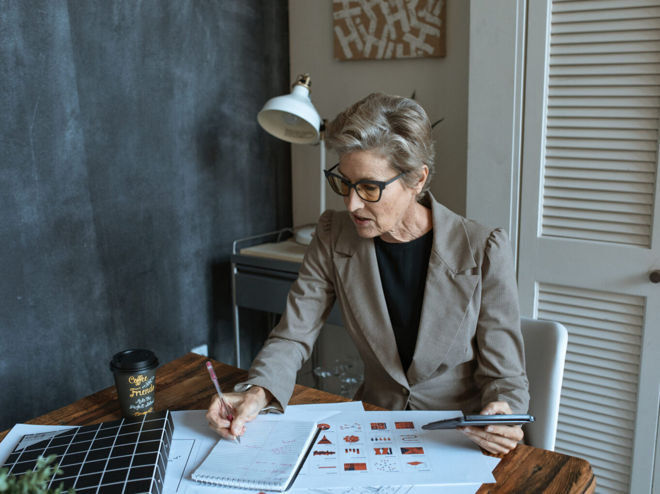 From above of concentrated senior female manager in formal outfit and eyeglasses taking notes in planner while working at table with various documents and takeaway coffee in cozy office