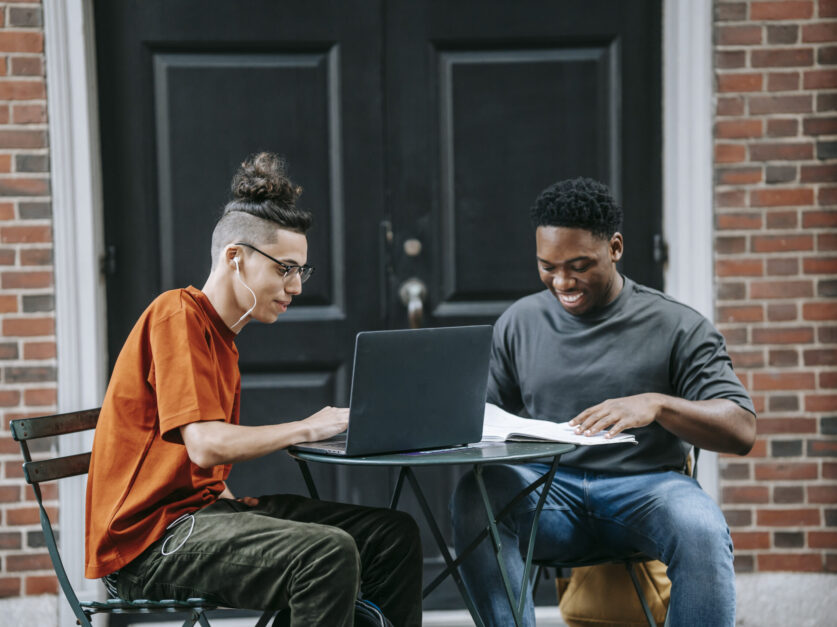 Positive young diverse male colleagues in casual clothes smiling and using laptop while working remotely on project sitting at table in street cafe